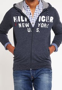 Felpa con zip colore navy, foderata in grigio, con grande scritta bianca "HILFIGER NEW YORK U.S.". Include tasche anteriori e cappuccio con cordino.