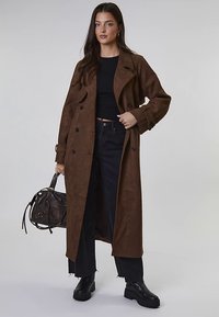 Brun långrock i mocka med dubbelknäppt design, skärpdetaljer vid ärmsluten och en avslappnad passform, kombinerad med svart topp och jeans.