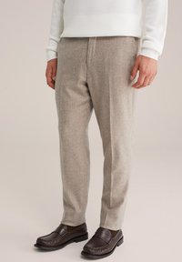 Mann in beigen Wollhosen, weißem Strickpullover und dunkelbraunen Lederloafers, der vor einem neutralen Hintergrund steht.