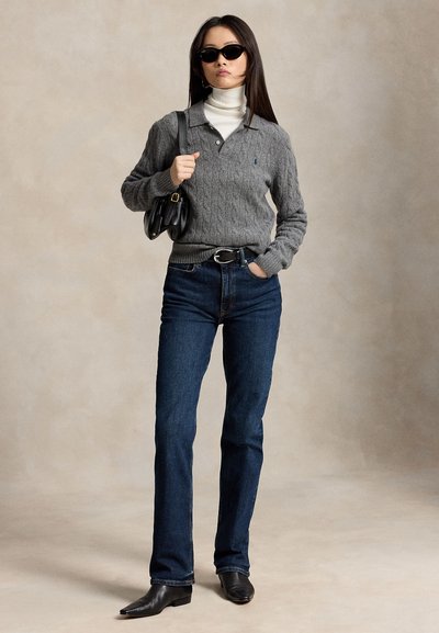 Femme portant des lunettes de soleil noires, un pull en maille torsadée gris sur un col roulé blanc, un jean bleu foncé, des bottes noires, et portant un sac à main noir à l'épaule.