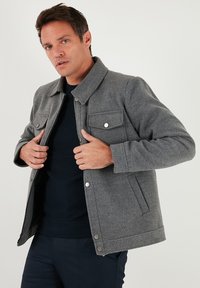 Jachetă gri din amestec de lână cu nasturi tip capse, două buzunare la piept și guler cu rever, purtată peste o cămașă neagră, afișată pe un fundal alb.