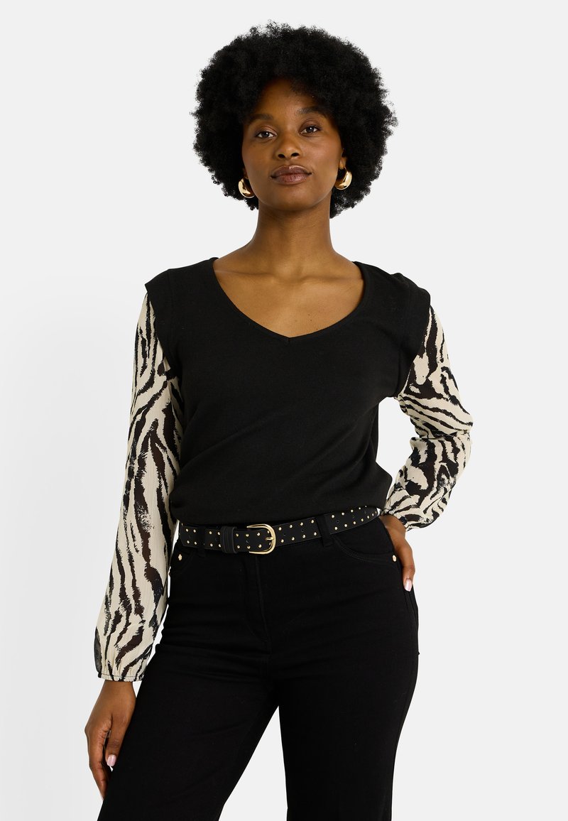 Top negro con escote en V, que presenta largas mangas de estampado animal en beige y negro. Combinado con pantalones negros de cintura alta y un cinturón con tachuelas.