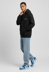 Haut à fermeture éclair noir avec logo en écriture blanche, associé à un jean bleu clair et des sneakers bleu/blanc, mettant en avant une coupe décontractée et un style casual.