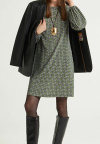 Robe verte à motifs géométriques, manches longues, associée à un blazer en cuir noir et des bottes noires matelassées jusqu'aux genoux. Collier pendentif en or.