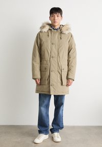 Khaki parka takki, jossa on turkista vuorattu huppu, etunapit, kaksi suurta taskua ja rento istuvuus, yhdistettynä sinisiin farkkuihin ja valkoisiin tennareihin.
