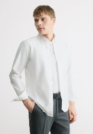 Jonge man die een wit overhemd draag over een wit T-shirt, grijze broek, met één hand in de zak, kijkt naar links tegen een effen achtergrond.