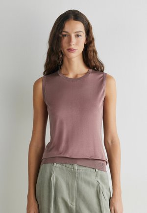 Junge Frau mit langen, welligen Haaren, die ein ärmelloses mauvefarbenes Top und hellgraue Hose trägt, steht vor einer schlichten weißen Wand.