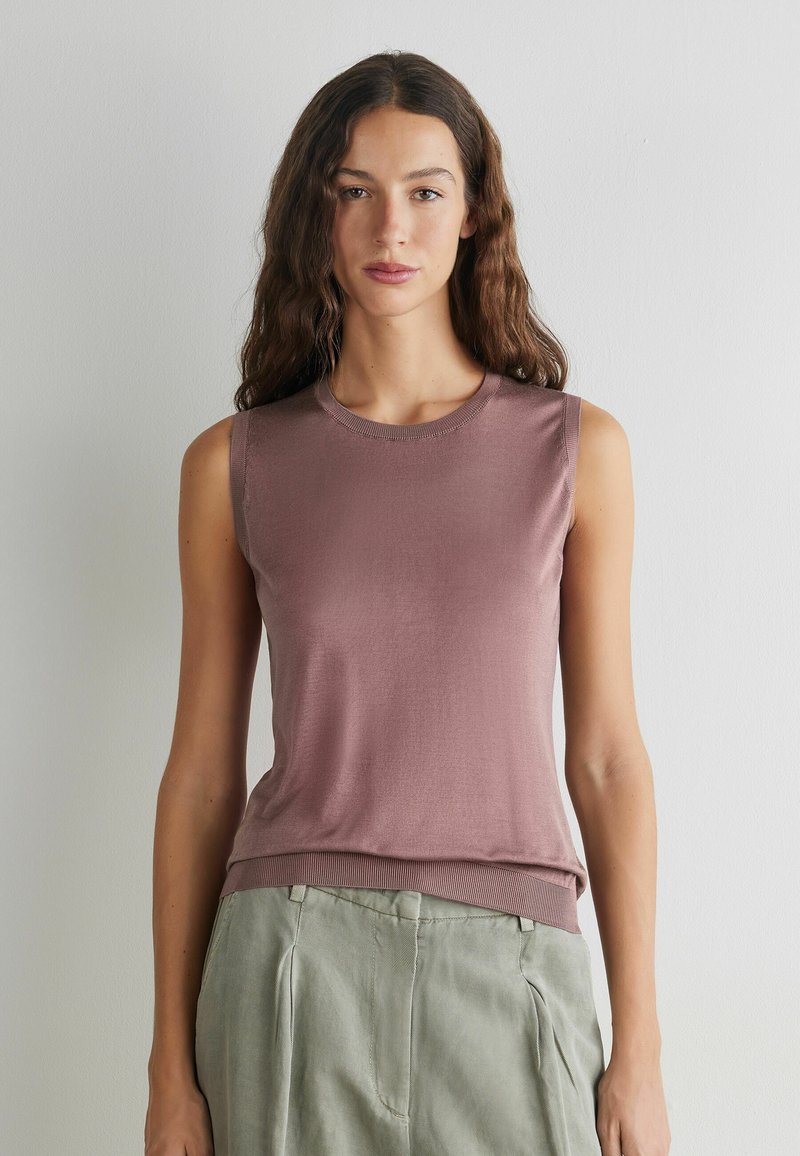 Young woman with long wavy hair wearing a sleeveless mauve top and light gray pants, standing against a plain white wall.