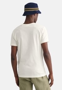 Hvid bomulds t-shirt med rund hals, vist på en person iført en marineblå bøttehat med grønne og gule striber samt lysegrønne bukser.