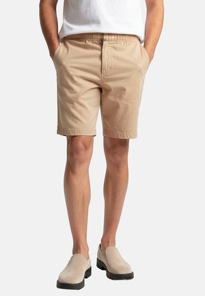 Homme portant un short beige, une chemise blanche rentrée, et des chaussures beige à enfiler, debout les mains dans les poches.