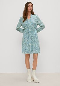 Abito azzurro chiaro con un motivo di sfumature verdi e blu. Presenta maniche lunghe, scollo a V e una vita elastica. Indossato con stivaletti bianchi alla caviglia.