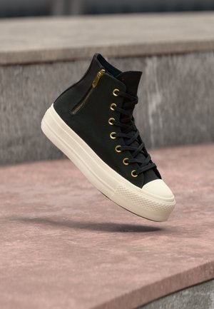 Zapatilla negra de caña alta con suela blanca y gruesa, puntera blanca, ojales dorados, cordones negros y una pequeña cremallera dorada en el lateral, flotando sobre el suelo.