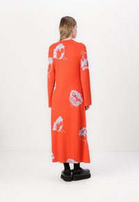 Abito lungo arancione in maglia con motivi floreali grigi, maniche lunghe e scollatura a girocollo, abbinato a scarpe con platform nere. Tessuto morbido.