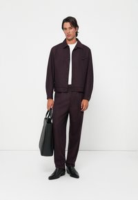 Man stands against white wall wearing dark purple suit, white shirt, black shoes, holding black leather tote bag in his left hand.