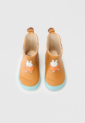 Bottes en caoutchouc marron à la cheville avec des semelles turquoise, présentant un design de lapin de dessin animé à l'avant. Texture lisse avec deux languettes de traction.