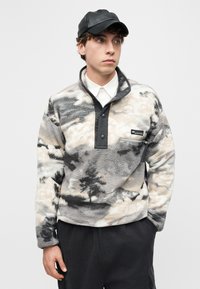 Νέος άνδρας που φοράει μαύρο καπέλο, λευκό πουκάμισο με γιακά και γκρι φλις πουλόβερ με ζιπ κι ανθεκτικ ά κουμπιά, με τα χέρια στις τσέπες του παντελονιού.