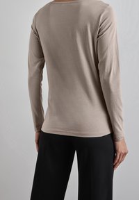 Beige långärmad topp i mjukt tyg, med rund halsringning och enkel sömnad. Parat med svarta byxor, med fokus på bakvy.