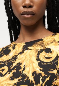 Κοντινή λήψη ενός ατόμου που φοράει τοπ με μαύρο και κίτρινο μοτίβο, με στροβιλισμένα σχέδια που μοιάζουν με πιτσιλίσματα και λεπτομέρειες με γραφή σε καλλιγραφικό στυλ.