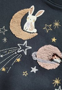 Lapin blanc assis sur un croissant de lune en paillettes pêche, entouré d'étoiles brodées et d'une planète pailletée sur un tissu foncé.