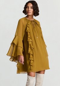 Senfgelbes Kleid mit Rüschärmeln, Schlitz am Ausschnitt und fließendem Design. Seidige Textur, kombiniert mit kniehohen weißen Stiefeln.