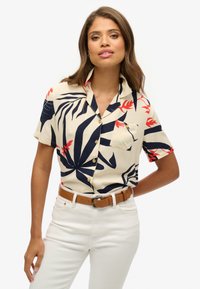 Camisa de manga corta en color crema con un patrón de hojas tropicales en azul marino y rojo, con botones en la parte delantera, un bolsillo en el pecho, combinada con jeans blancos y un cinturón marrón.