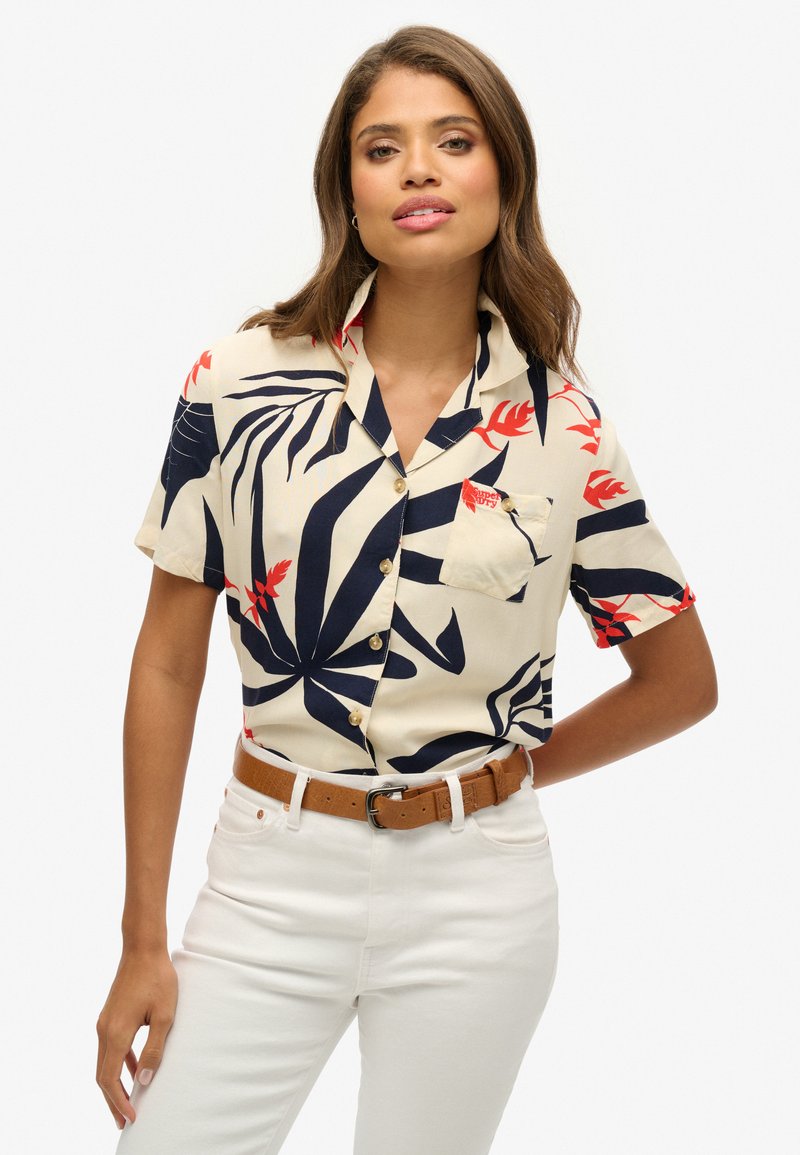 Korte mouw crèmekleurig shirt met marineblauw en rood tropisch bladenpatroon, knoopsluiting aan de voorkant, enkele borstzak, gedragen met witte jeans en een bruine riem.