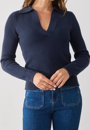 Kobieta ubrana w granatowy prążkowany sweter z długim rękawem z kołnierzem i dekoltem w serek, w parze z niebieskimi dżinsami, z rękami splecionymi na talii.