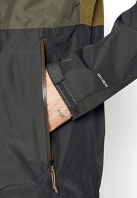 Main partiellement dans la poche zippée d'une veste DRYVENT noire et olive, montrant un tatouage au poignet avec le mot "TIME".