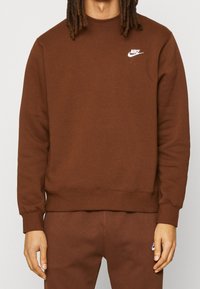 Person wearing brown Nike sweatshirt and matching brown pants, with Nike logos visible on the chest and thigh.