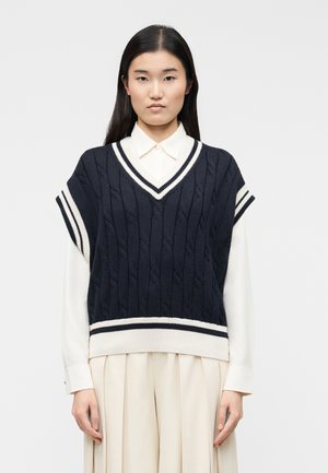 Fiatal nő hosszú, sötét hajjal, sötétkék kábelmintás ujjatlan pulóvert visel fehér gombos ing fölött, bézs pliszírozott szoknyával.