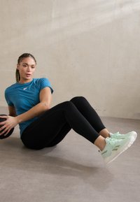 Mentazöld sportcipő fehér részletekkel, fekete leggings-szel és türkiz, testhez álló pólóval párosítva, edzés környezetében.