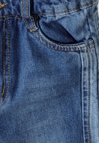 Tejido de denim en azul con costuras amarillas contrastantes. Presenta una cinturilla elástica y un bolsillo lateral. Textura suave con pliegues sutiles.