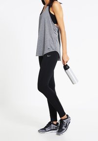 Grå ärmlös tanktop med djupa sidor, svarta leggings och svart-vita mönstrade sneakers, hållande en silverfärgad vattenflaska.