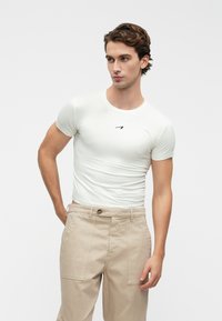 Pieguļošs balts t-krekls ar īsām piedurknēm un apaļu izgriezumu, ar mazu melnu logotipu uz krūtīm, apvienots ar bēšiem biksēm.