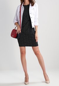 Black polka dot dress with a fitted shape, paired with a white denim jacket. Accented with a red crossbody bag and nude heels.