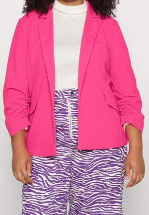 Person iført en skarp pink blazer over en hvid top og lilla og hvide zebra print bukser med lynlås foran.