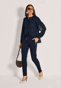 Marinebluse mit faltigen Ärmeln, passenden taillierten Hosen und beigen spitzen Absätzen. Accessoire: kleine dunkle Handtasche mit Metallnieten.