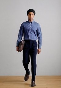 Mėlyna susegama marškiniai, aptemptos tamsios kelnės, rudi odiniai batai ir ruda rankinė ant peties. Modelis pristatytas ant vienspalvio fono.