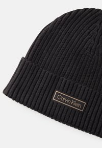 Czarna dziana czapka beanie z prążkowaną teksturą, złożonym mankietem i prostokątną metalową łatą z logo marki w białej czcionce.