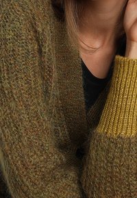 Lähikuva henkilöstä, jolla on päällään teksturoitu ruskea neuletakki, jossa on keltainen hiha mustan topin päällä, näyttäen kaulan ja osan kasvoista.