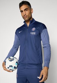 Marineblå treningsjakke med glidelås og lysere blå ermer, med en logo på venstre bryst og en glidelåslomme, holdt av en person med en fotball.