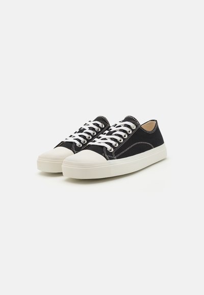 Svarta canvas sneakers med vita snören, en rundad vit gummikappa på tån och kontrasterande vit sömnad på sidorna. Lågt profildesign.