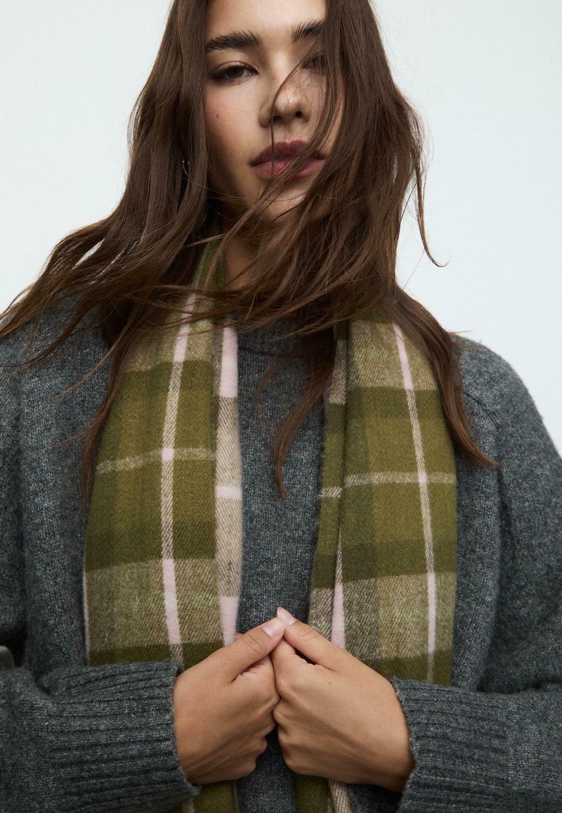 Écharpe à carreaux olive et beige avec des accents roses, portée sur un pull gris texturé, sur fond neutre.
