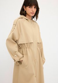 Beige, långärmad kappa med huva, elastisk midja, framknäppning och sidofickor; slät textur; har detaljer i guldfärgade metall.