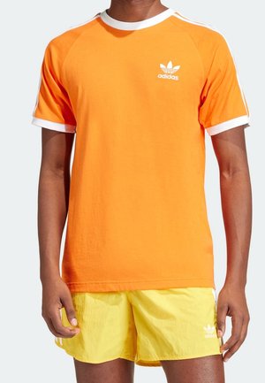 Homme portant un t-shirt Adidas orange vif avec des bordures blanches et un short Adidas jaune avec des bandes blanches sur les côtés.