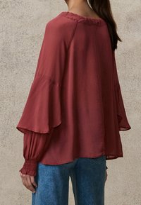 Blusa bordeaux in tessuto leggero con lunghe maniche a campana, polsini arricciati e vestibilità ampia; indossata con jeans blu.