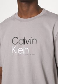 Gray cotton t-shirt featuring a crew neck and printed logo with "Calvin" in black and "Klein" in white, along with additional text beneath.