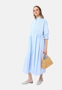 Robe rayée bleu clair avec un devant boutonné, des manches longues et un corsage ajusté. Associée à des chaussures plates grises et un sac en osier.