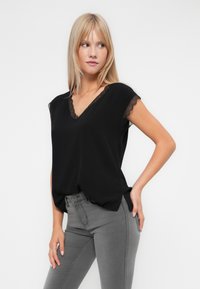 Blusa negra de manga corta con escote en V y detalles de encaje en el escote y las mangas, hecha de una tela suave. Llevada con jeans ajustados grises.