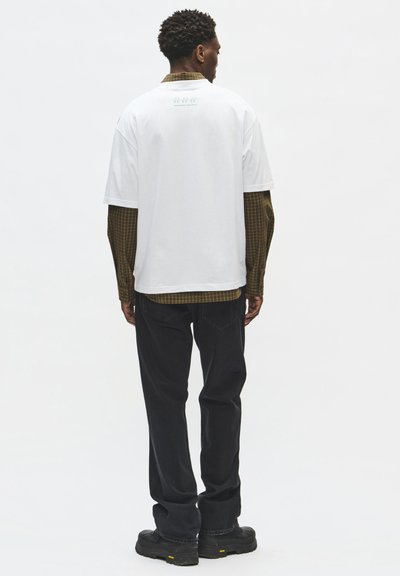 Homme portant une chemise blanche à manches courtes par-dessus une chemise marron à carreaux à manches longues, pantalon noir et chaussures noires, debout de dos.